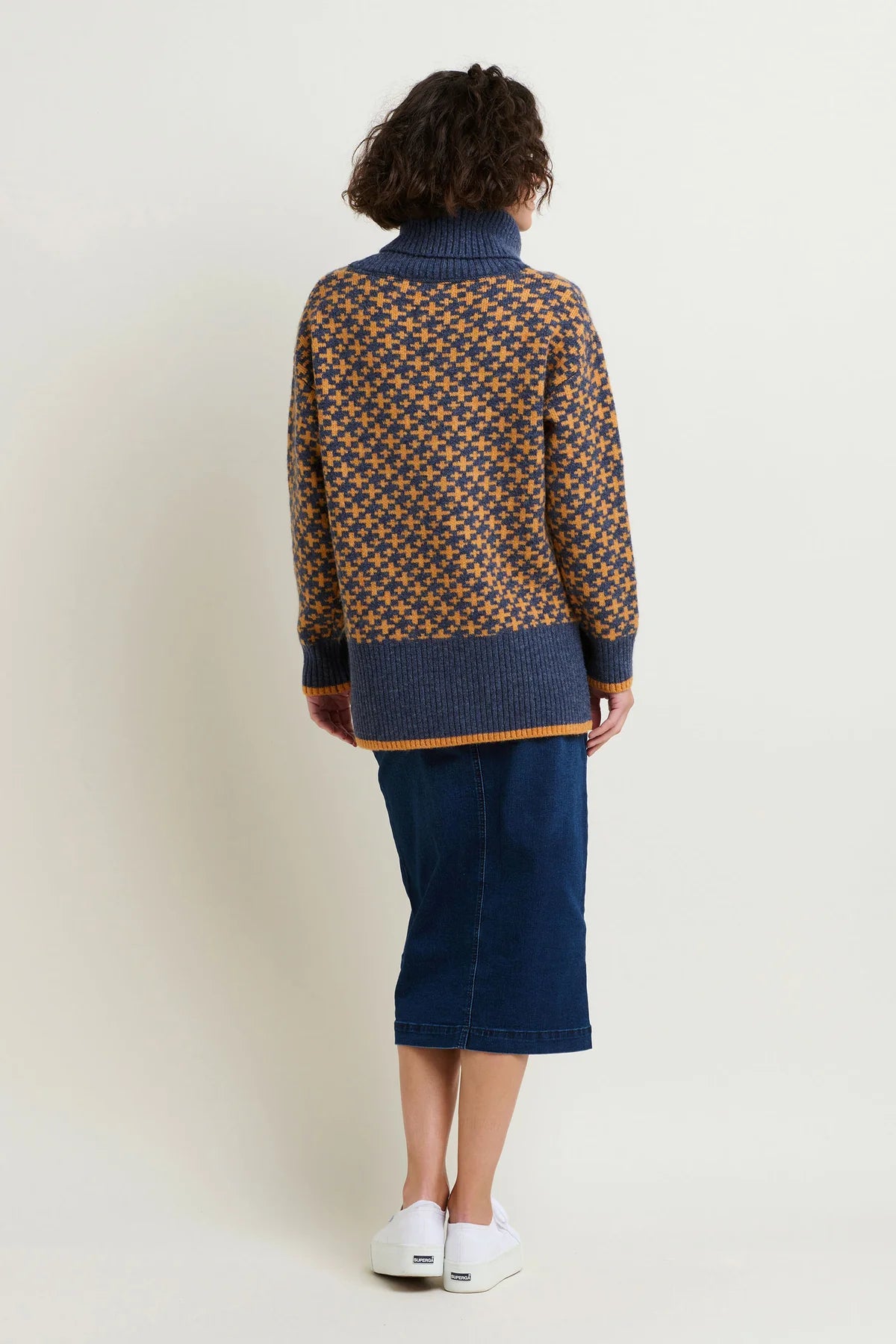 Back view of a woman wearing a knitted jumper with blue loose roll neck, blue main fabric with small and slightly larger crosses knitted through. Worn with a dark blue denim skirt.